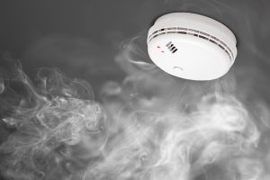 A ceiling-mounted smoke detector with a red light is surrounded by wisps of smoke against a dark background.